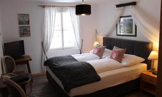 A cozy bedroom with a double bed and soft lighting. Decorative pictures hang on the wall, and a window lets in plenty of daylight.