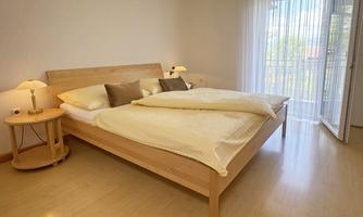 A bright bedroom with a large double bed and wooden furniture. The window offers a view of a balcony and lets in plenty of daylight.