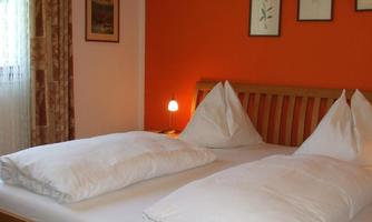 A cozy bedroom with a wooden bed and white pillows. The walls are painted in warm orange, and there is soft light from a lamp.