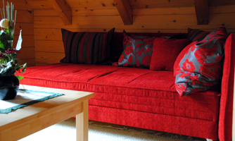 A red sofa with colorful cushions is in a cozy room. A wooden table and a small plant complement the decor.