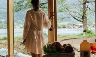 A person stands in front of a large window front and looks out at an idyllic lake. In the foreground, there is a bowl of fresh fruit.
