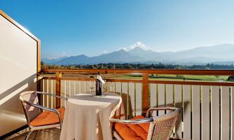 A balcony with a table and two chairs offers a beautiful view of the mountains. A bottle and a glass are on the table, and the sun shines brightly in the sky.