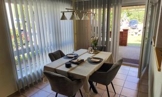 A bright dining area with a table and elegant chairs. Large windows with blinds provide a view of the outdoors.
