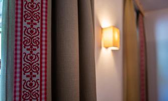 A room with curtains in beige and a striped border in red and white. In the background, a wall lamp is installed.