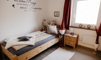 A cozy bedroom with a wooden bed and decorative pillows. An inspirational quote is on the wall, and the room has a warm, inviting atmosphere.