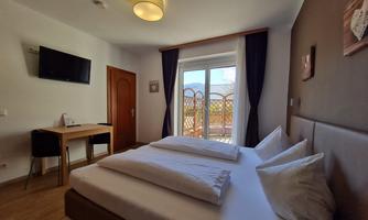A cozy hotel room with a double bed, a table, and chairs. Large windows provide daylight and a view outside.