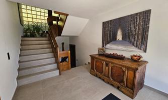 A bright entrance area with a staircase and a wooden piece of furniture. On the wall hangs a large picture of a forested path.