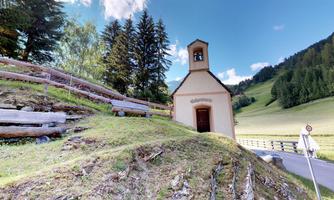 A small church stands on a hill, surrounded by trees and meadows. In the background, gentle hills and a clear sky can be seen.