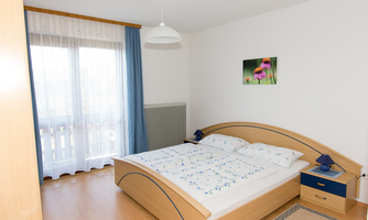 A bright bedroom with a double bed and a blue dresser. Bright curtains hang in front of the windows, and a picture of flowers adorns the wall.