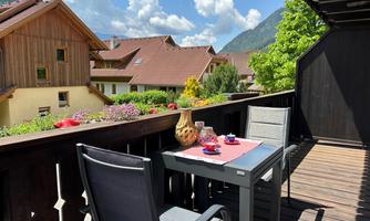 A cozy balcony with a table and two chairs. In the background, green mountains and picturesque houses can be seen.