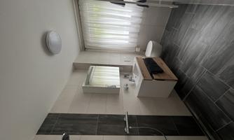 A modern bathroom with a shower cubicle, a sink, and a large window. The floor is tiled with dark tiles and the walls are light.