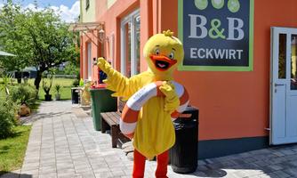 A cheerful yellow duck stands in front of the B&B Eckwirt. It holds a lifebuoy in its hand and smiles.