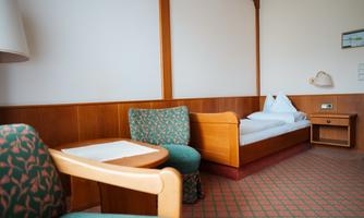 A simple room with a single bed, a table, and an armchair. The walls are bright and the decor is modern.