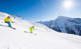 Two skiers are riding down a snow-covered slope in the mountains. The sky is blue and the sun is shining brightly.
