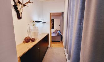 A narrow hallway with a wooden shelf and decorative elements. On the right side, there is a view of a cozy living room.