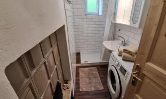A modern bathroom with a shower, a sink, and a washing machine. The walls are covered in light-colored tiles, and the window lets in natural light.