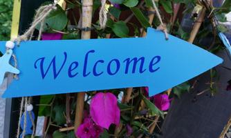 A blue welcome sign with the word "Welcome" stands next to colorful bougainvillea blooms. The sign points to the right in an arrow shape.