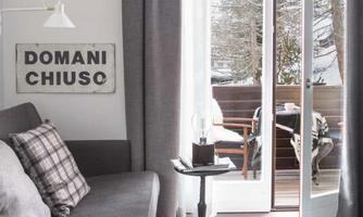A modern living room with a cozy sofa corner and a small table. The large window front leads to a balcony with a view of nature.