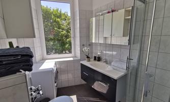 A modern bathroom with a shower, a sink, and a washing machine. The window lets in a lot of daylight and offers a view of green trees.