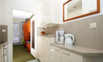 A modern kitchen with bright cabinets and a kettle on the countertop. In the background, an open space with a rug and a window can be seen.