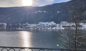A picturesque winter view of a lake, surrounded by snow-covered mountains. The sun shines brightly over the scene and reflects in the water.