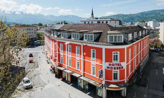 A red hotel building named "Hotel Mössner" stands at a street intersection. In the background, mountains and a blue sky can be seen.
