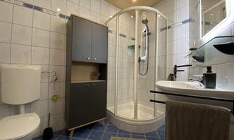 A modern bathroom with a shower, a sink, and a toilet. The walls are tiled, and there is a simple cabinet for storage.