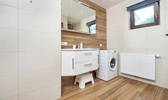 A modern bathroom with a white sink and a mirror. On the right side, there is a washing machine, and the floor is fitted with wooden floorboards.