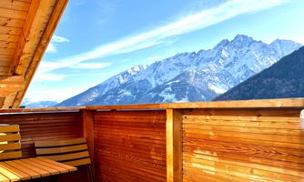 A balcony with wooden paneling and two chairs. In the background, majestic snow-covered mountains and a clear blue sky can be seen.