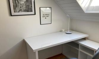 A bright workspace with a white desk and a blue chair. Two pictures are hanging on the wall, while a lamp sits on the table.
