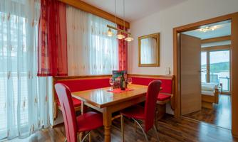 A cozy dining area with a wooden table and red chairs. The windows are decorated with light curtains and offer a view of the scenery.