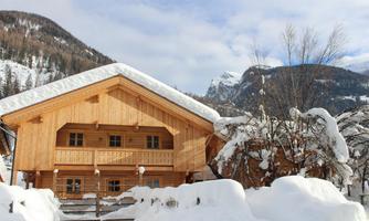 A cozy wooden house in the snow, surrounded by mountains. The sky is partially cloudy and the landscape looks wintry.