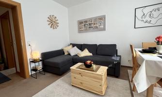 A cozy living room with a gray sofa and a wooden table. There are decorative elements on the walls and a dining area.