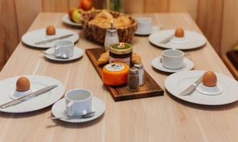 A beautifully set breakfast table with eggs, cups, and fresh rolls. In the middle, there is cheese and jam in a jar.