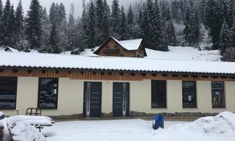 A modern building in the snow, surrounded by tall fir trees. The landscape is wintry and peaceful.