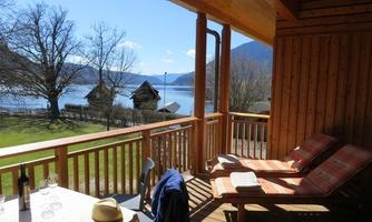 A cozy balcony with two loungers and a table on which a bottle of wine and glasses are placed. In the background, a lake and mountains are visible under a blue sky and sunshine.