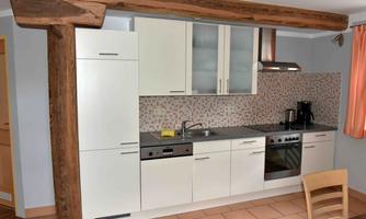 A modern kitchen with white cabinets and a shiny countertop. The room has a wooden pillar and a stylish tiled wall.