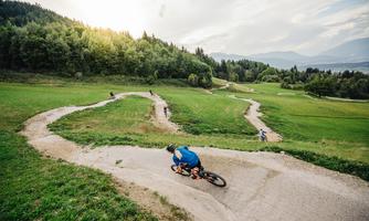 A biker rides on a winding path through a green landscape. In the background, more cyclists and forests can be seen.