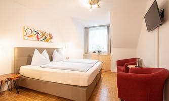 A cozy bedroom with a double bed and two red armchairs. The room has a television and a window with a view outside.