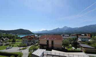 A picturesque landscape with mountains and a clear blue sky. In the foreground, there are houses and a green surrounding.