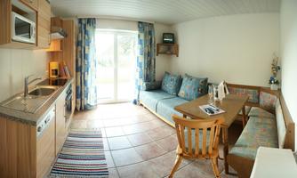 A cozy holiday apartment with a kitchenette and a dining area. The windows let in plenty of daylight and the room is cheerfully decorated.