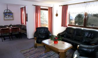 A cozy living room with black leather furniture and a wooden table. Large windows let in plenty of daylight and offer a view of the surroundings.