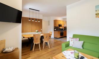 A modern living and dining room with a green sofa and a wooden table. The kitchen in the background is well-equipped and bright.