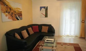 A cozy living room with a black couch and colorful cushions. The room has a large window with curtains and some artworks on the walls.