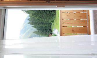 A view from an open window onto a green meadow and trees in the background. The balcony is made of wood and the curtains are slightly open.
