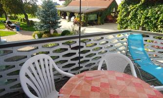 A cozy balcony with a round table and two chairs. In the background, green plants and part of the property are visible.