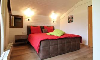 A cozy bedroom with a large red bed and colorful cushions. Bright walls and a wooden floor create a warm atmosphere.