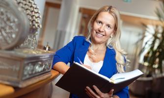A smiling woman in a blue blazer is standing at a table, holding a notebook in her hand. In the background, plants and modern interior are visible.
