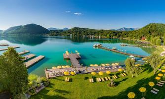 A picturesque lake landscape with crystal blue water and green hills in the background. Sun loungers line the shore and a pier leads into the water.
