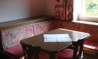 A cozy dining area with a table and chairs. A cross hangs on the wall, and bright light falls through the window.
